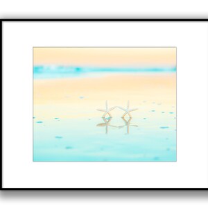May include: Two white starfish on a sandy beach with turquoise water reflecting the sky. The sky is a gradient of yellow and blue.