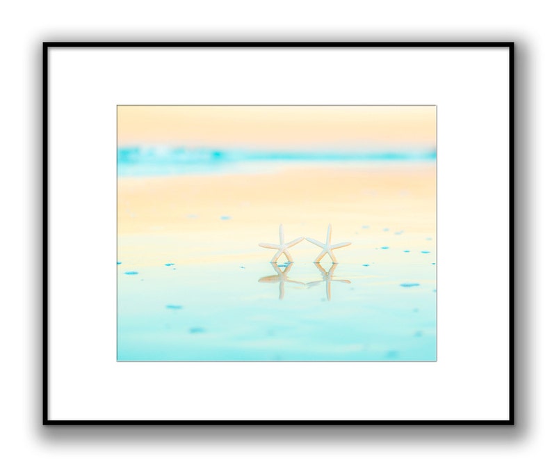 May include: Two white starfish on a sandy beach with turquoise water reflecting the sky. The sky is a gradient of yellow and blue.