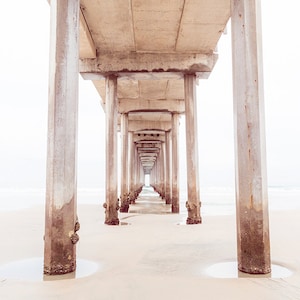 May include: A view of a concrete pier extending out over a sandy beach. The pier is made up of a series of columns that create a tunnel-like effect.