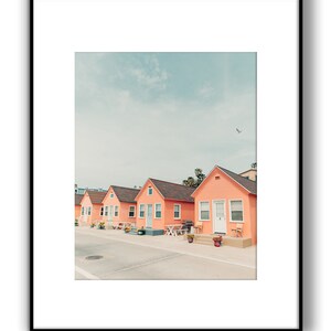 May include: A row of five pastel pink beach houses with white trim and white doors. The houses are lined up on a street with a light gray asphalt surface. The sky is a light blue with a few white clouds.
