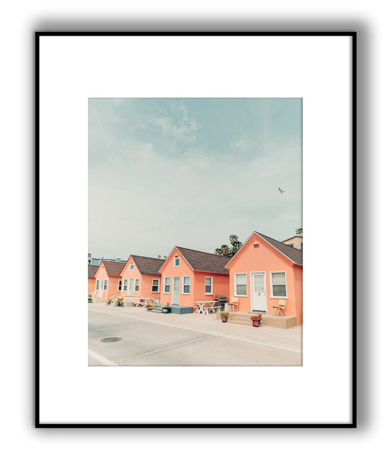 May include: A row of five pastel pink beach houses with white trim and white doors. The houses are lined up on a street with a light gray asphalt surface. The sky is a light blue with a few white clouds.