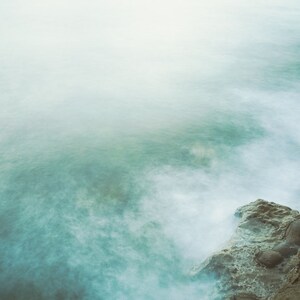 May include: A misty, ethereal seascape with a rocky shoreline. The water is a soft, muted blue-green, and the rocks are a dark brown. The image has a dreamy, atmospheric quality.