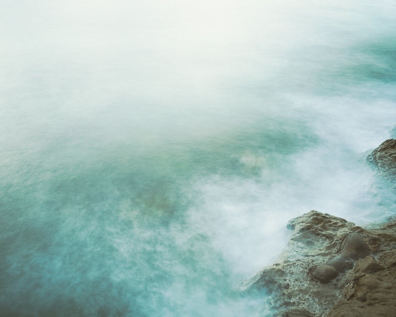May include: A misty, ethereal seascape with a rocky shoreline. The water is a soft, muted blue-green, and the rocks are a dark brown. The image has a dreamy, atmospheric quality.
