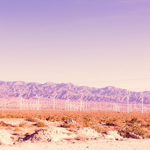 May include: A field of wind turbines in a desert landscape with mountains in the background. The sky is a pale pink color.