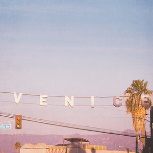 May include: A white sign with the word "VENICE" spelled out in large letters hangs from power lines against a pale pink sky. A palm tree stands in the foreground.