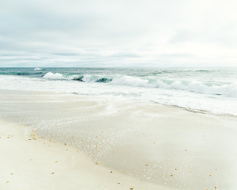May include: A white sandy beach with foamy waves crashing on the shore. The sky is overcast and the water is a light blue.