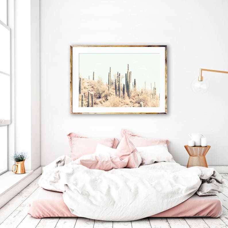 Pu&ograve; includere: Una stampa incorniciata di un paesaggio desertico con cactus nei toni del beige e del marrone &egrave; appesa sopra un letto con biancheria da letto rosa e bianca. La stanza ha un'estetica minimalista con un tavolino dorato e una finestra.