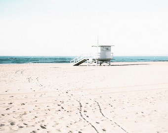 May include: A white lifeguard stand with the number 115 on the side, on a sandy beach with footprints leading away from it. The ocean is in the background.
