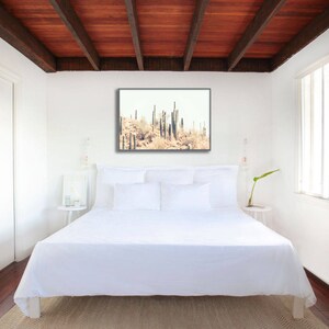 Pu&ograve; includere: Camera da letto con letto bianco, cuscini e biancheria da letto. Una stampa incorniciata di cactus &egrave; appesa sopra il letto. La stanza ha un soffitto in legno e un tappeto colorato neutro.