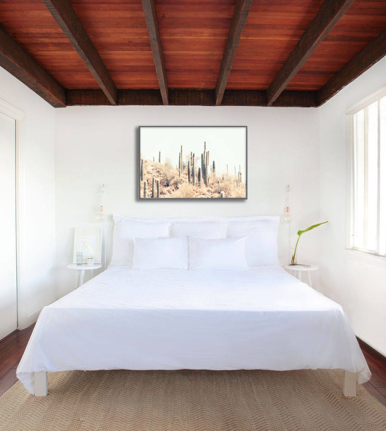 Pu&ograve; includere: Camera da letto con letto bianco, cuscini e biancheria da letto. Una stampa incorniciata di cactus &egrave; appesa sopra il letto. La stanza ha un soffitto in legno e un tappeto colorato neutro.