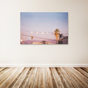 May include: A photograph of a pink and purple sunset over the city of Venice, California. The iconic "Venice" sign is strung across a street with power lines. A palm tree is visible in the background.