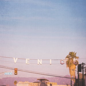 May include: A white sign with the word "VENICE" spelled out in large letters hangs from power lines against a pastel pink and blue sky. A palm tree stands in the foreground.