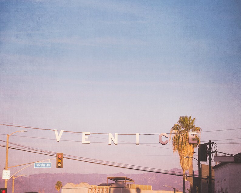 May include: A white sign with the word "VENICE" spelled out in large letters hangs from power lines against a pastel pink and blue sky. A palm tree stands in the foreground.
