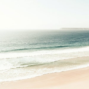 May include: A panoramic view of a sandy beach with white foamy waves crashing on the shore. The ocean is a deep blue, and a distant rocky coastline is visible in the background.