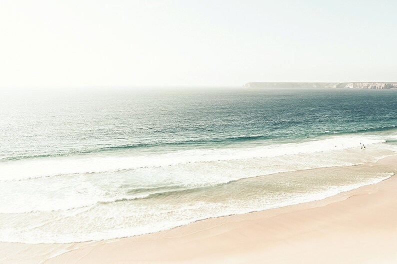 May include: A panoramic view of a sandy beach with white foamy waves crashing on the shore. The ocean is a deep blue, and a distant rocky coastline is visible in the background.