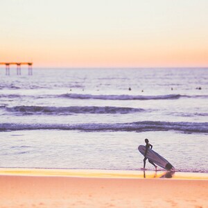 May include: A person walks along a sandy beach carrying a surfboard. The ocean is in the background with a pier in the distance. The sky is a soft pink and purple.