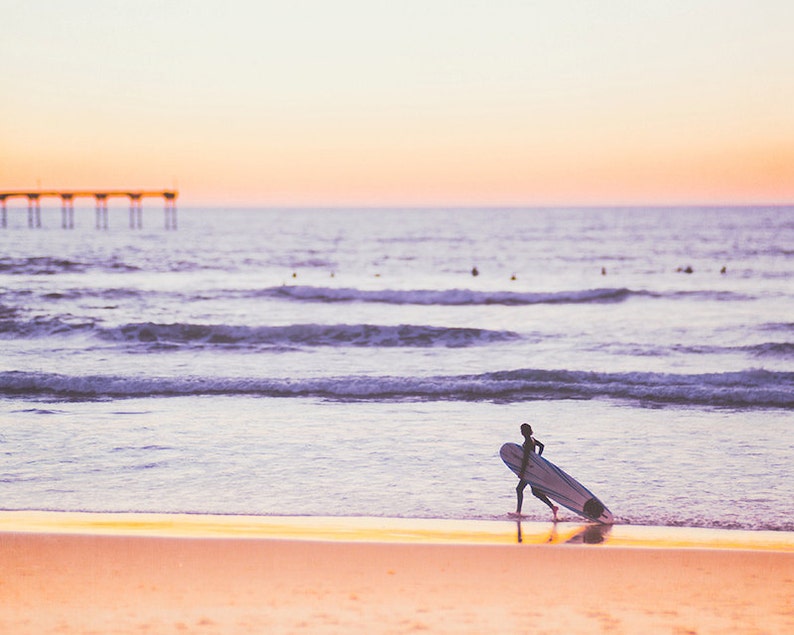May include: A person walks along a sandy beach carrying a surfboard. The ocean is in the background with a pier in the distance. The sky is a soft pink and purple.