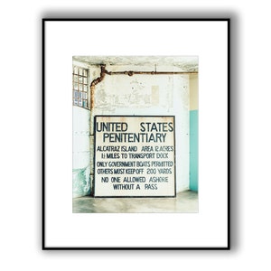 May include: A black and white sign with the text "UNITED STATES PENITENTIARY" and details about Alcatraz Island. The sign is mounted on a white wall with peeling paint.