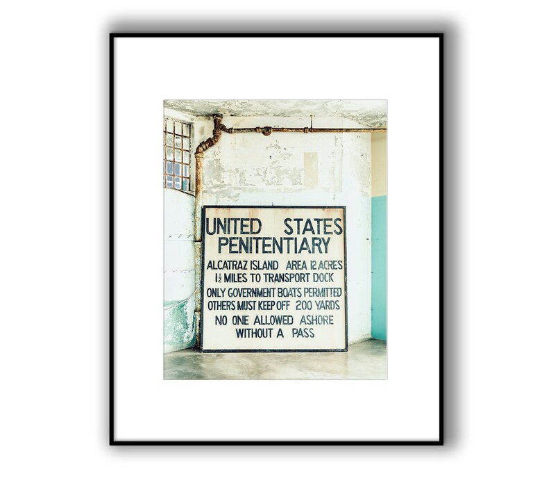 May include: A black and white sign with the text "UNITED STATES PENITENTIARY" and details about Alcatraz Island. The sign is mounted on a white wall with peeling paint.