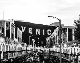 May include: Black and white photo of a street in Venice, California. The street is lined with shops and restaurants, and the word "VENICE" is displayed in large letters on a building.
