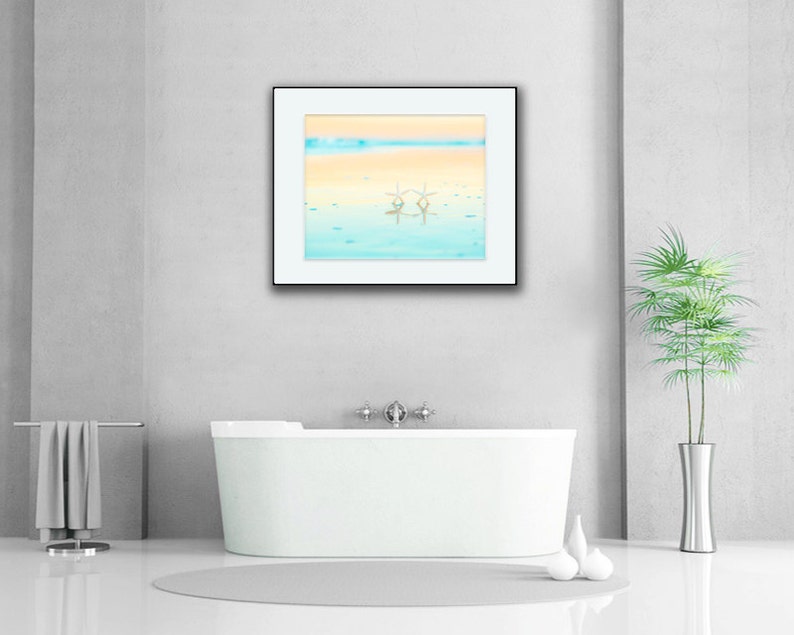 May include: A white bathtub with a framed picture of a beach scene above it. The picture shows a blue and yellow sky with two white starfish in the water.