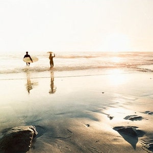 May include: Two people walk out of the ocean carrying surfboards on a sunny day. The beach is sandy and the water is clear.