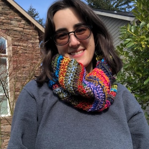 May include: A person wearing a gray sweatshirt with a colorful crocheted infinity scarf. The scarf is made with a variety of colors, including red, orange, yellow, green, blue, and purple.