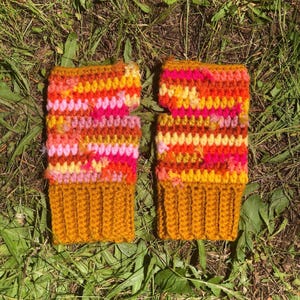 Peut inclure: Une paire de mitaines faites à la main aux couleurs vives. Les mitaines présentent un poignet côtelé jaune moutarde et un corps de mailles crochetées orange, rose et jaune mélangées. Les mitaines sont posées sur de l'herbe verte.