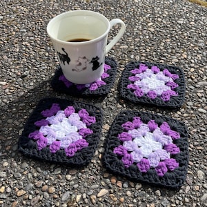 May include: A white mug with black cat designs, filled with coffee, sits on a black, purple, and white crocheted coaster. Three more coasters with the same granny square pattern are also visible.