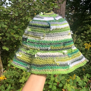 May include: A handmade crochet bucket hat in shades of green, white, and gray. The hat features a wide brim and a textured, striped pattern. The hat is held up against a backdrop of green foliage.