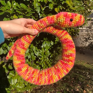 May include: A crocheted snake toy in a bright pink, yellow, and green color pattern. The snake is curled into a circle and has black eyes.