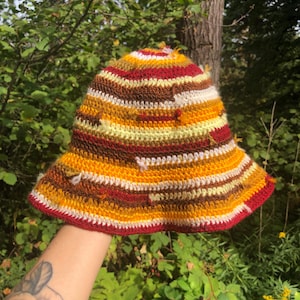 May include: A handmade crochet bucket hat with a colorful striped pattern. The hat features bands of red, yellow, brown, and white. The brim is trimmed in red. The hat is held up by a person.