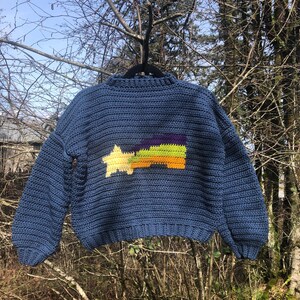 May include: A blue crochet sweater with a star and rainbow design. The sweater features a crew neck and long sleeves. The rainbow design includes yellow, orange, green, and purple colors. The sweater is hanging on a black hanger against a natural background.