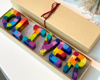 May include: A set of multi-coloured wax crayons shaped into the letters that spell the name OLIVER. The crayons feature a marbled pattern of blue, green, yellow, orange, purple, and red. They are arranged inside a rectangular cardboard gift box.