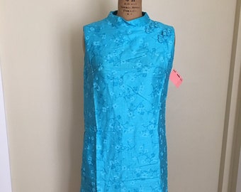 Könnte beinhalten: Ein ärmelloses türkisfarbenes Kleid mit weißem Blumenmuster. Das Kleid hat einen Stehkragen und einen Knopfverschluss am Hals. Das Kleid befindet sich auf einer Kleiderpuppe.