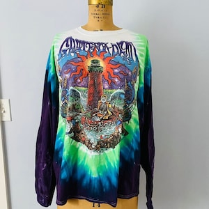 May include: A tie-dye long-sleeve shirt with a colorful graphic of a lighthouse, a skeleton in a boat, and a sun with a starry sky. The shirt is purple, green, and blue.