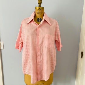May include: A pink striped short-sleeved button-down shirt with a pocket on the left side. The shirt is on a dress form.
