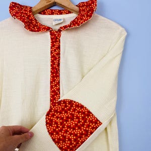 May include: Cream-colored waffle-knit robe with red floral accents. The robe features a ruffled collar, a red floral stripe down the front, and red floral elbow patches. The robe is hanging on a wooden hanger.