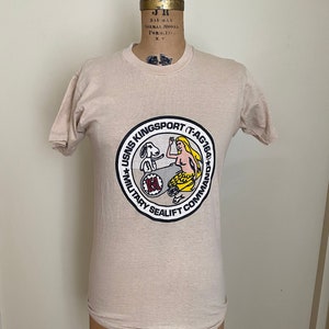 May include: A tan crew neck t-shirt featuring a circular emblem with the text "USNS Kingsport (T-AG164) Military Sealift Command." The emblem depicts a mermaid, a dog, and other nautical elements. The shirt is displayed on a mannequin.