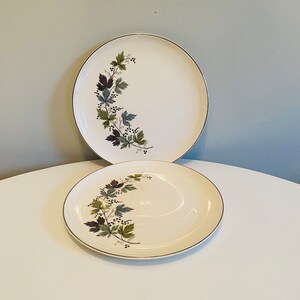 Op de afbeelding: Twee witte keramische borden met een gouden rand en een rankontwerp met groene bladeren en zwarte bessen.