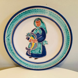 Puede incluir: Un plato de cerámica blanco con un borde azul y verde. El centro del plato presenta una ilustración pintada a mano de una mujer con un vestido azul y una chal verde que lleva una cesta de pescado.
