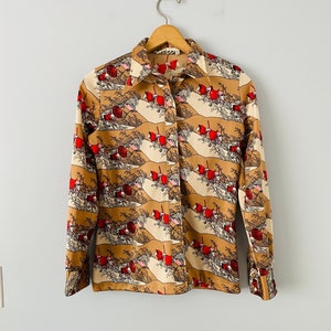 May include: A button-down shirt with a repeating pattern of red figures and landscape elements on a tan and cream background. The shirt has long sleeves and a collared neckline. The shirt is hanging on a wooden hanger.