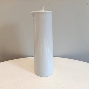 May include: A tall, white ceramic jug with a lid and a spout. The jug has a cylindrical body with vertical ribbed detailing on the lower half. The jug is on a white surface.