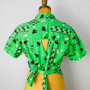 Puede incluir: Una camisa verde brillante con mangas cortas, con un patr&oacute;n repetido de siluetas de animales negros, p&aacute;jaros blancos y formas geom&eacute;tricas. La camisa est&aacute; atada a la cintura y se muestra en un maniqu&iacute;.