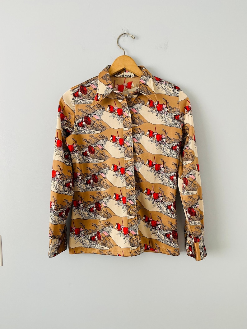 May include: A button-down shirt with a repeating pattern of red figures and landscape elements on a tan and cream background. The shirt has long sleeves and a collared neckline. The shirt is hanging on a wooden hanger.