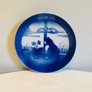 Könnte beinhalten: Blau-weiße Zierteller mit einer schwarz-weißen Katze an einem Fenster mit einer Winterszene. Der Teller hat einen blauen Rand mit strukturiertem Design und das Jahr "1970" ist auf dem Teller aufgedruckt.