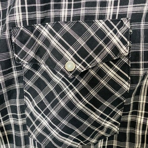 May include: A black and white plaid shirt with a flap pocket on the left side. The pocket has a diagonal plaid pattern and a silver button.
