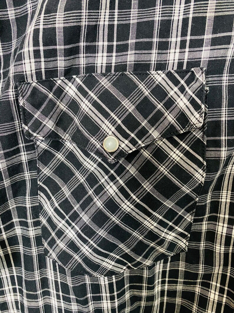 May include: A black and white plaid shirt with a flap pocket on the left side. The pocket has a diagonal plaid pattern and a silver button.