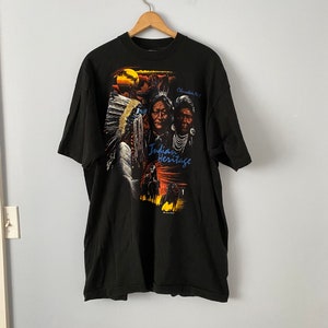 Puede incluir: Camiseta negra con un gráfico colorido de figuras nativas americanas y la frase "Indian Heritage". El diseño incluye retratos, una puesta de sol y siluetas de jinetes. La camiseta está colgada de una percha de madera.