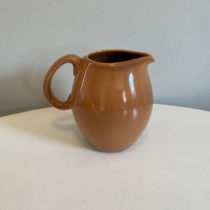 May include: A brown ceramic jug with a rounded body and a handle. The jug is sitting on a white surface.